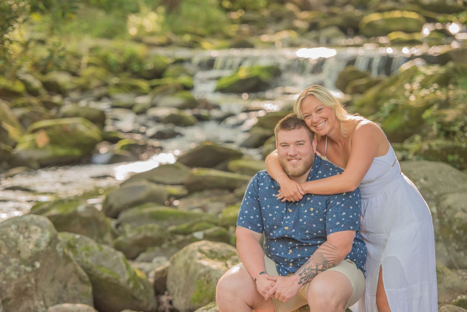 A n engagement couple poses together on mossy rocks near a cascading waterfall on a sunny afternoon.