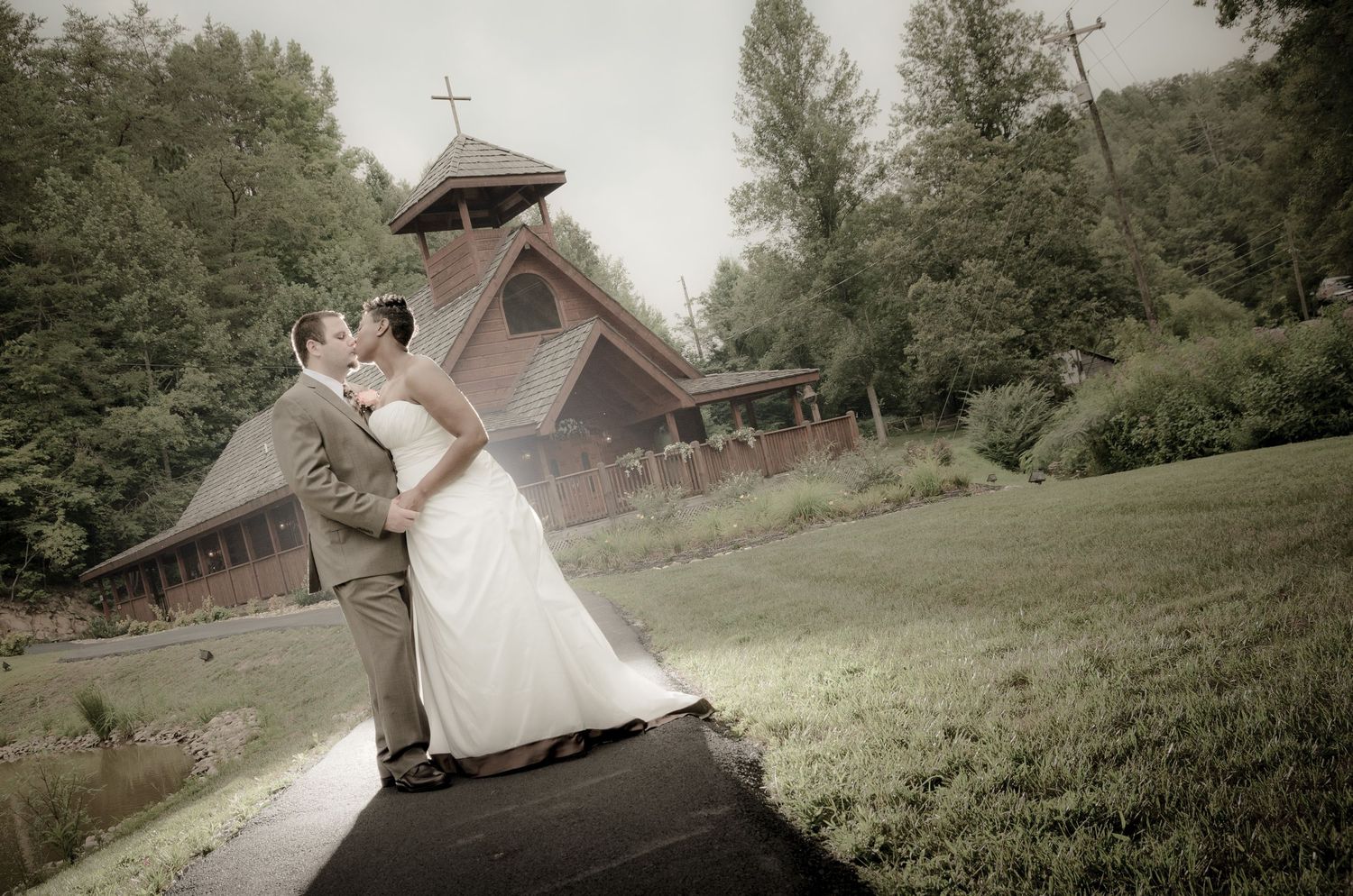 A couple embraces on a path in front of a rustic wooden chapel surrounded by lush green trees in Sevierville TN.