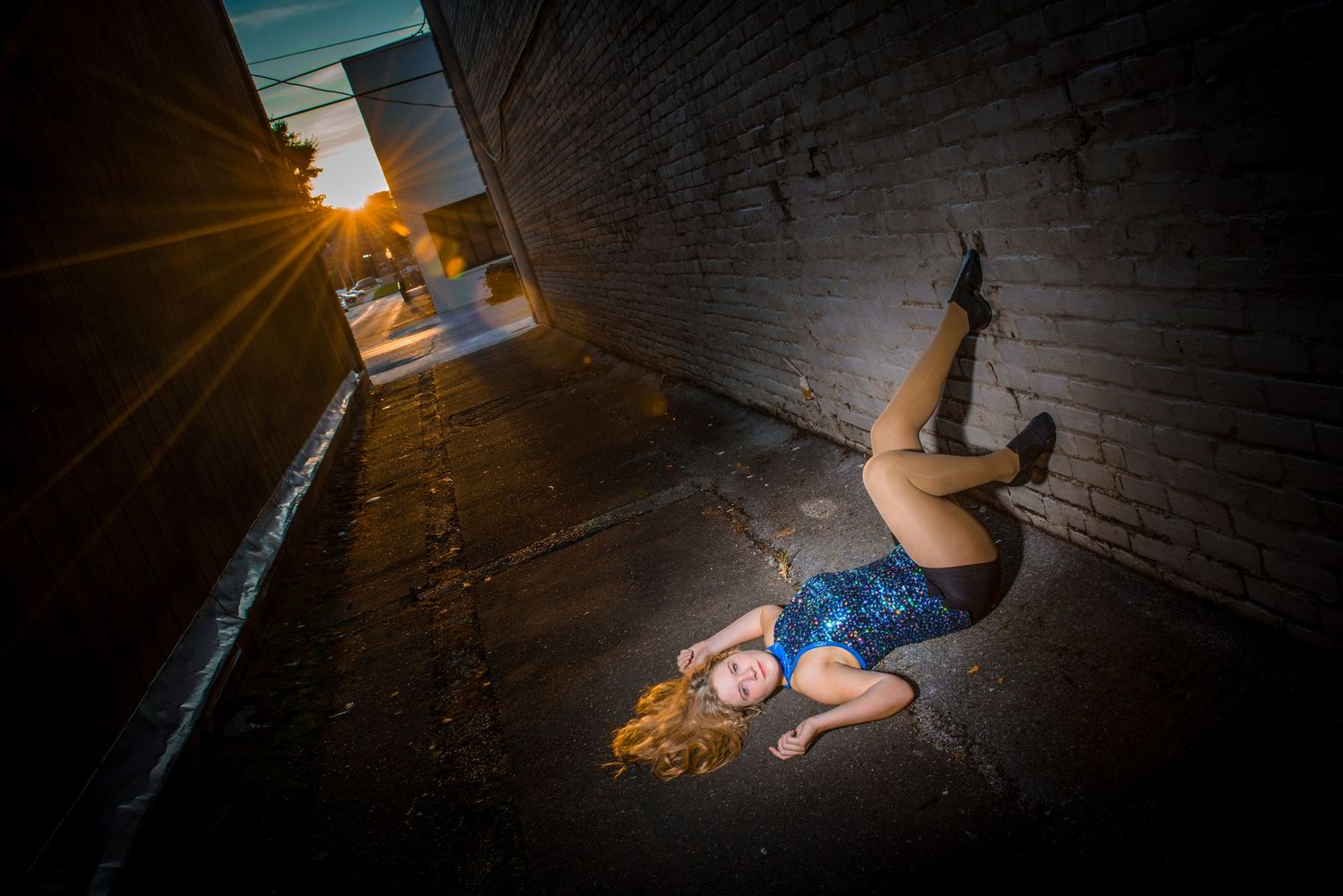 A person in athletic wear lies on the ground in a dramatic pose as sunlight streams through a narrow alleyway.
