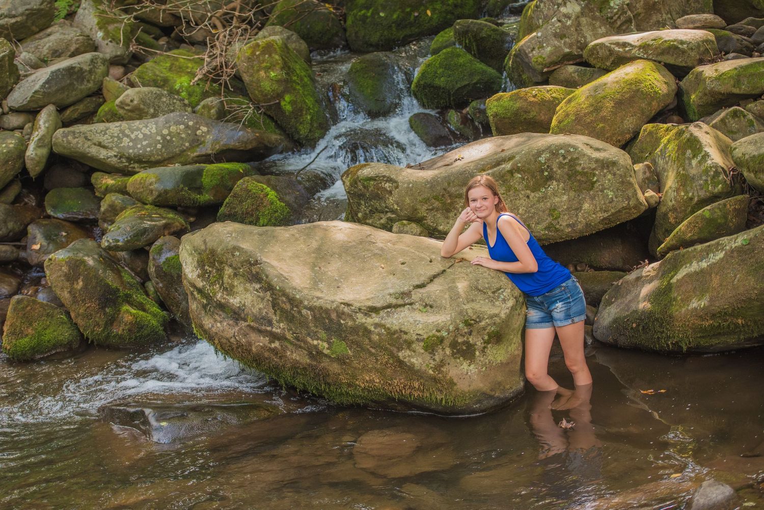 A person in blue stands by a mossy rock formation with cascading water flowing in the background at a mountain stream.