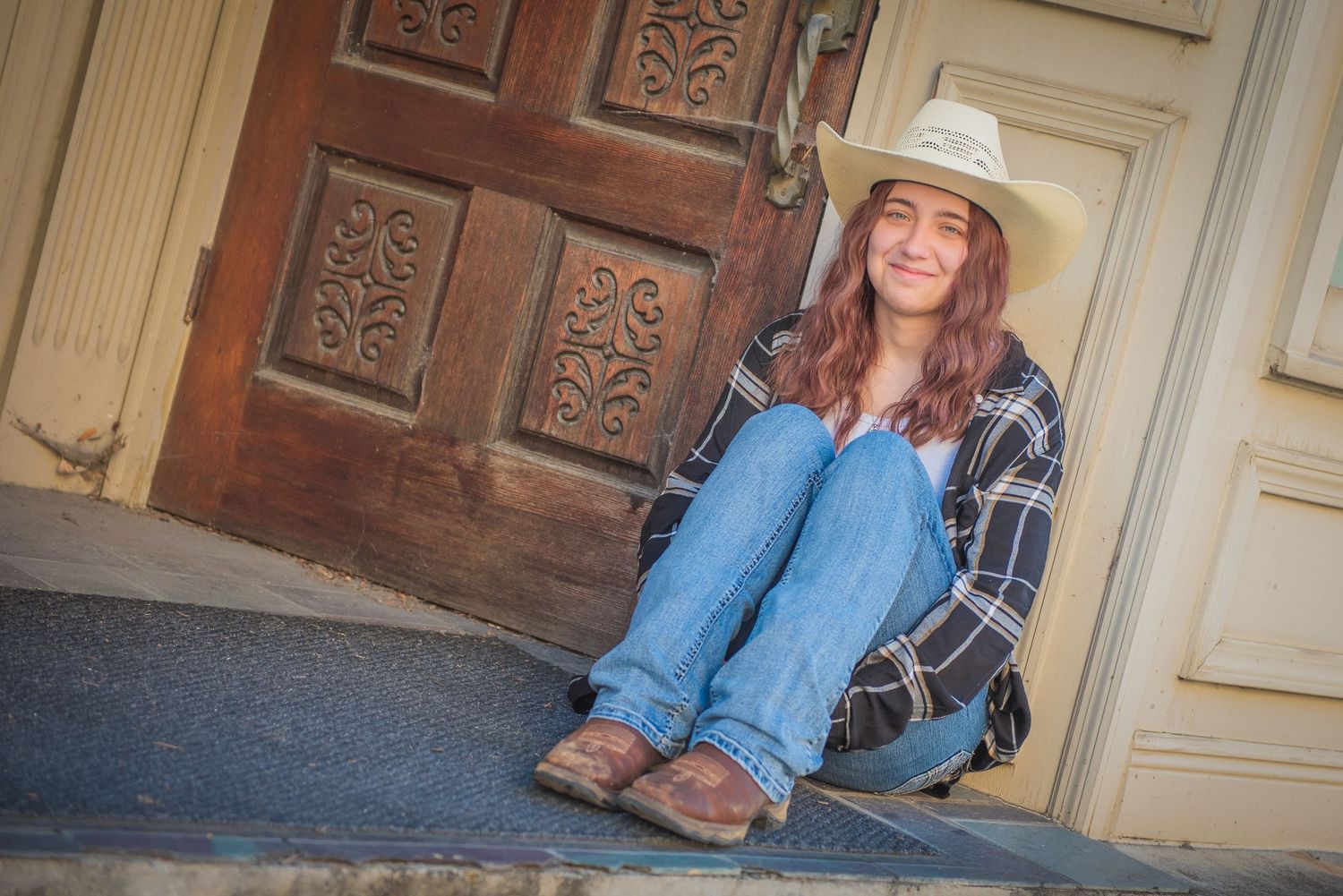 Someone wearing a cowboy hat, plaid shirt and jeans sits casually on stone steps in front of an ornate wooden door.