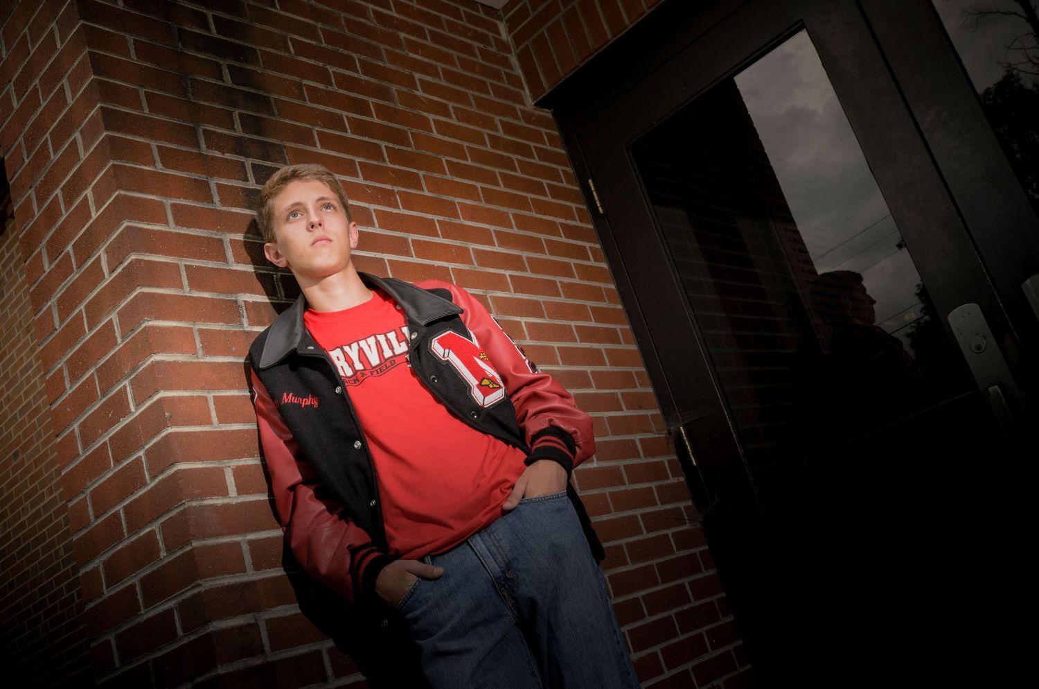 Person wearing a red sweatshirt leans against brick wall in dramatic lighting with dark shadows.