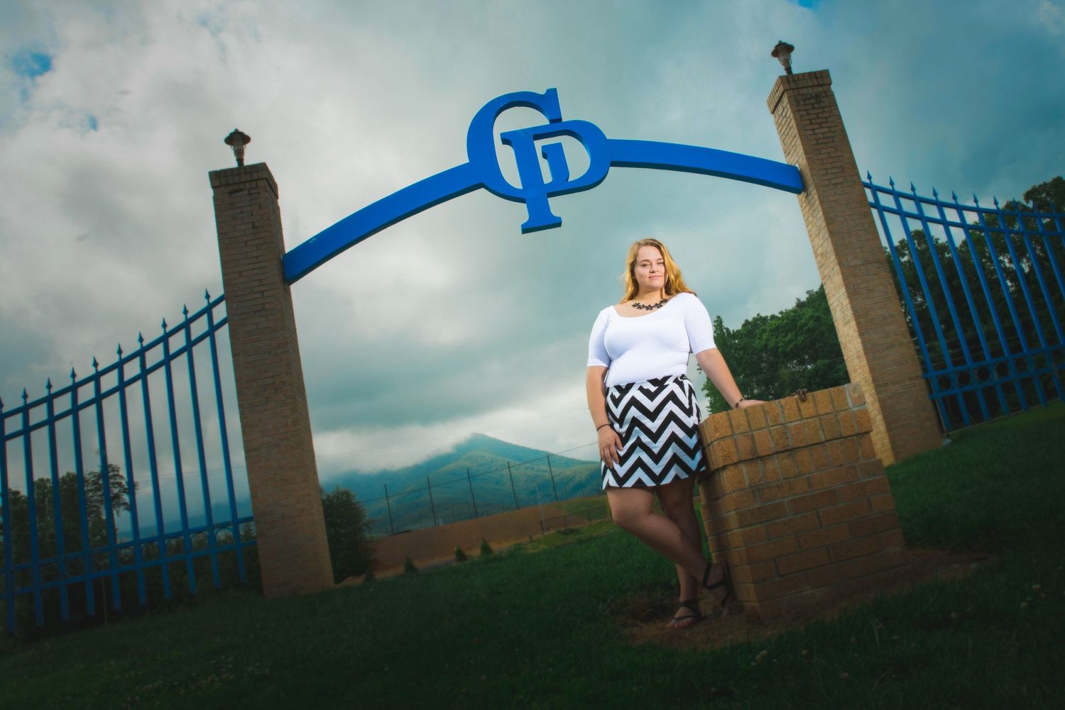 Someone stands near large decorative blue gates framed by stone pillars with mountains and cloudy sky in the background.
