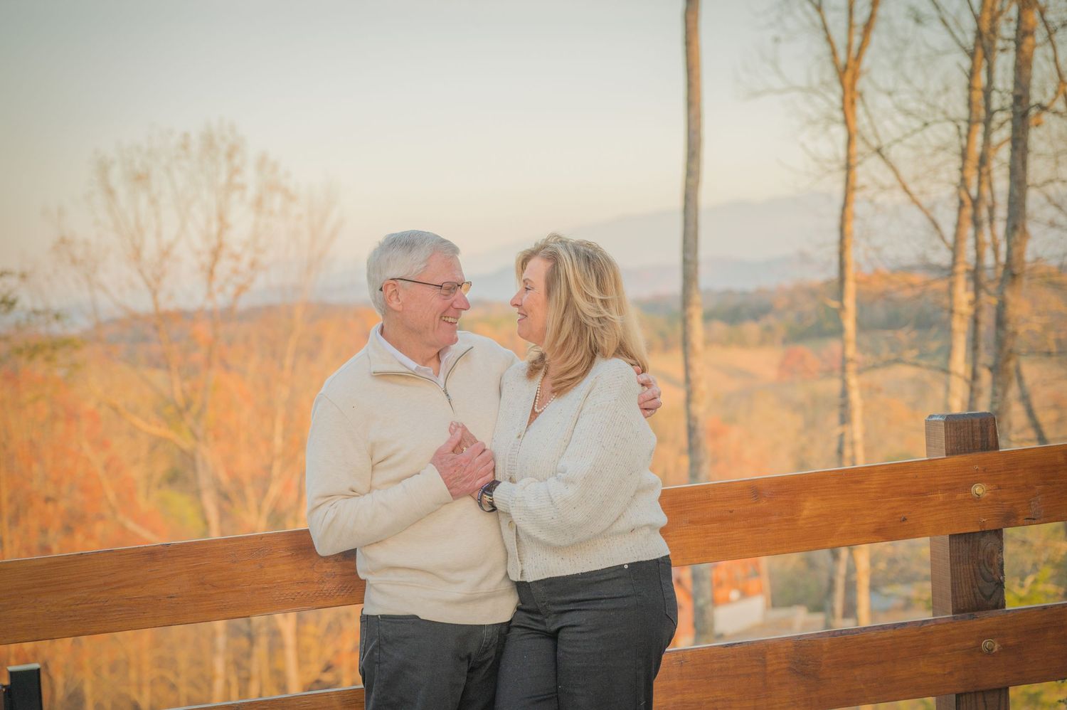 A couple in matching white sweaters share a tender moment on a wooden deck overlooking an autumn landscape at sunset in Pigeon Forge.