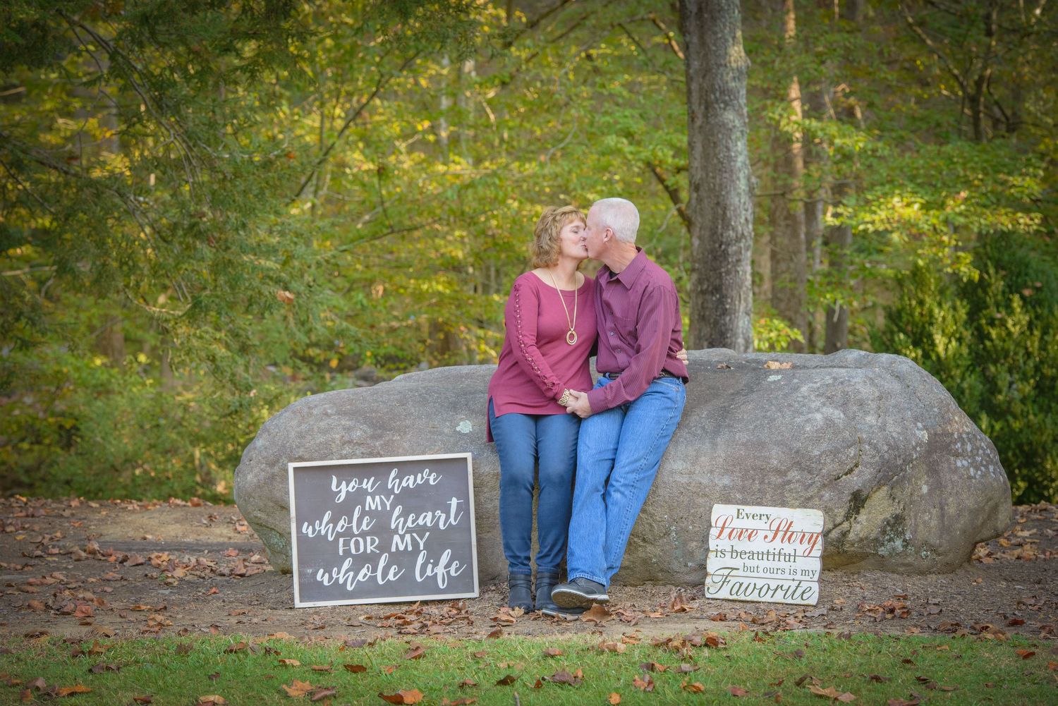 A couple sits together on a large rock in an autumn forest setting with two decorative signs beside them by Gatlinburg Photographers TN.