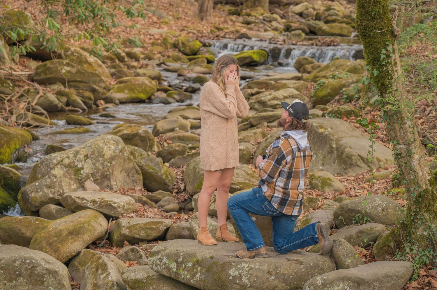 A serene proposal moment captured on mossy rocks beside a cascading waterfall in an autumn forest setting by Sevierville TN Photographers.