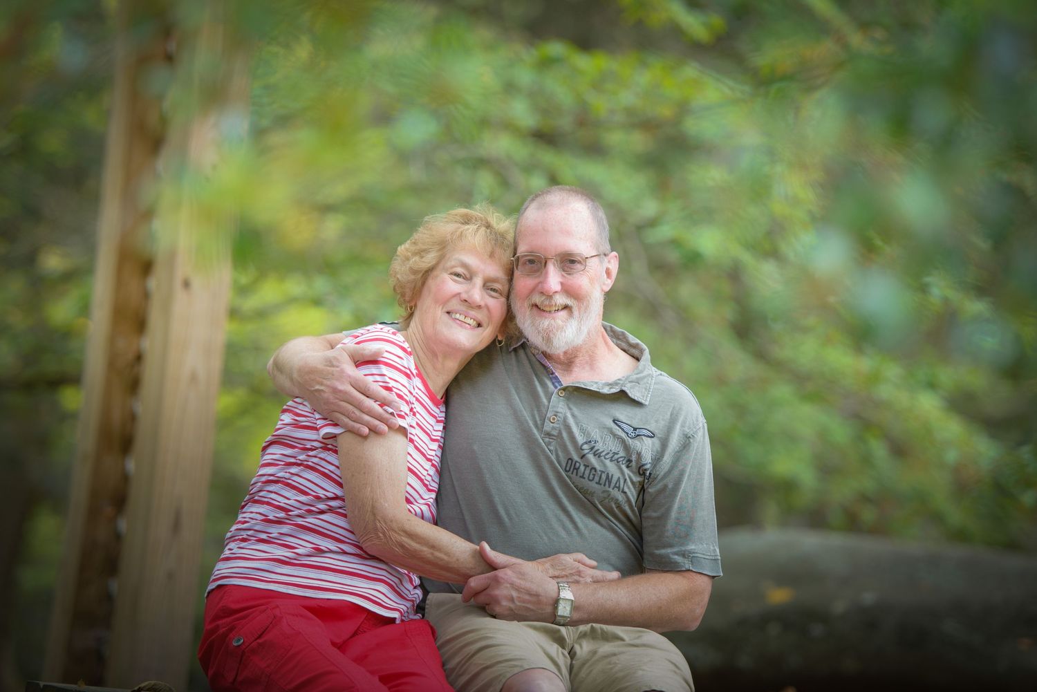 A couple sits close together on a wooden bench surrounded by soft green foliage in an outdoor setting by Sevierville TN Photographers.