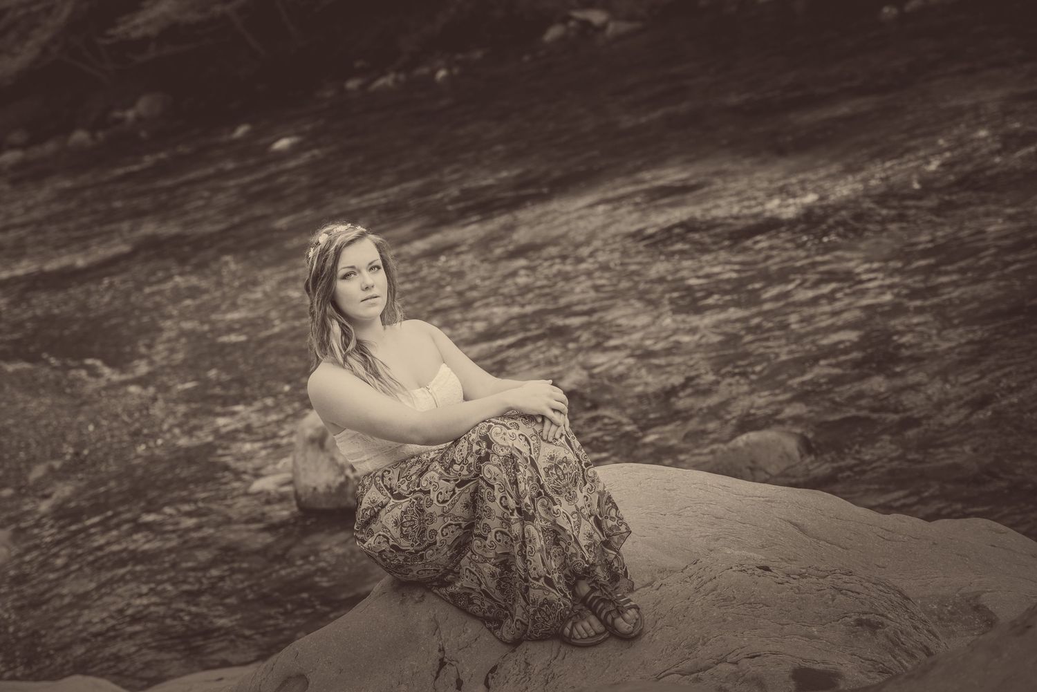 A vintage sepia-toned artistic portrait of a person resting by a rocky shoreline in a flowing dress.