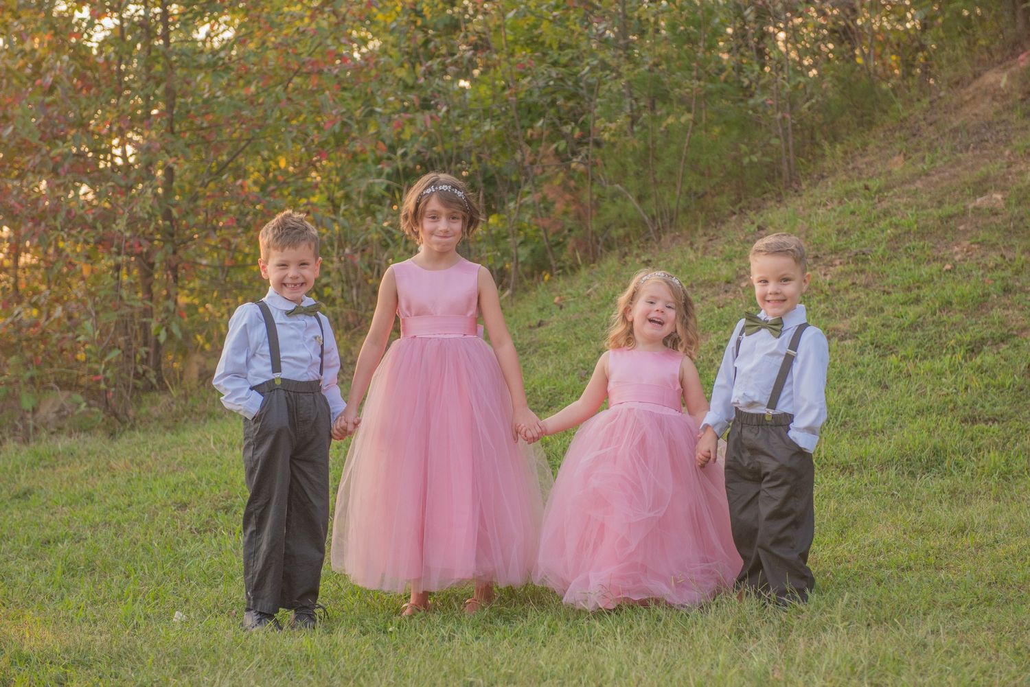 Children in formal attire including pink tulle dresses and navy suspenders pose together on a grassy hillside.