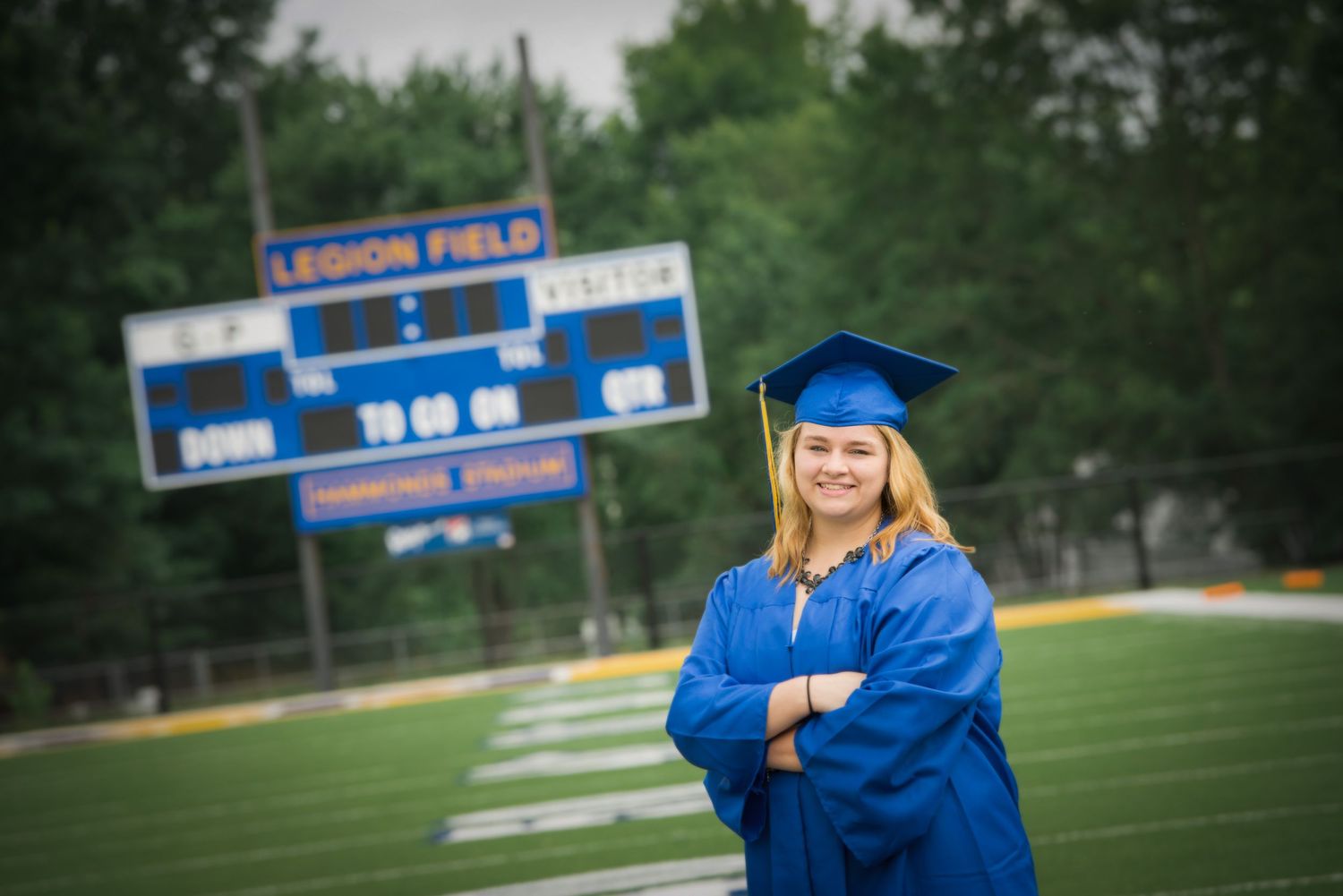 Graduate in blue cap and gown poses confidently on football field with scoreboard visible in background.