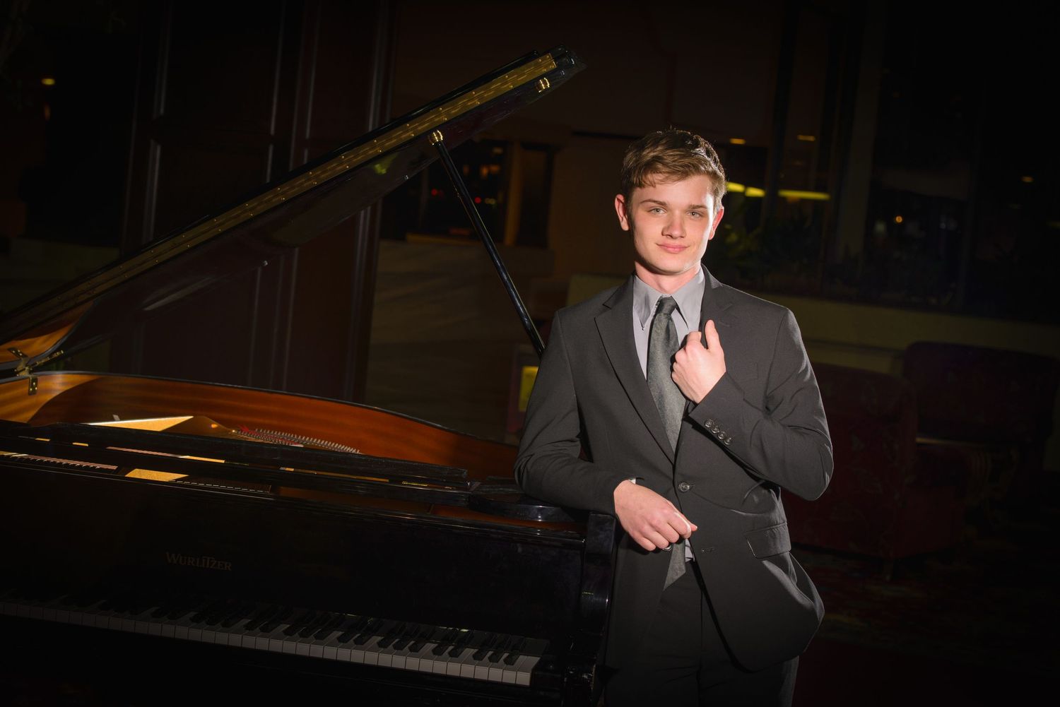 A performer in a gray suit stands next to a grand piano on a dimly lit stage.