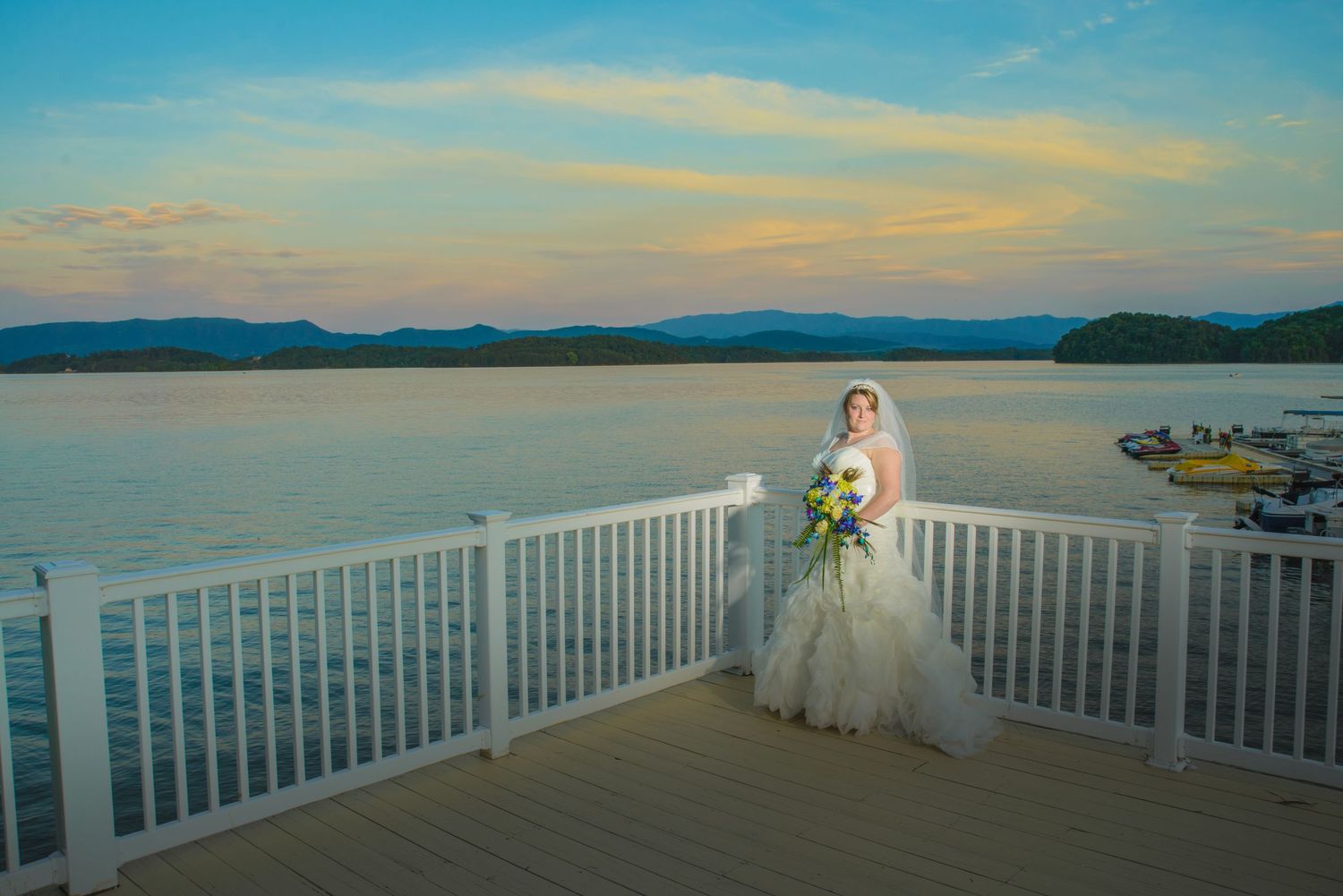 Dandridge Wedding Photographers portrait of a bride in a white gown stands on a waterfront deck overlooking a serene lake and mountains.