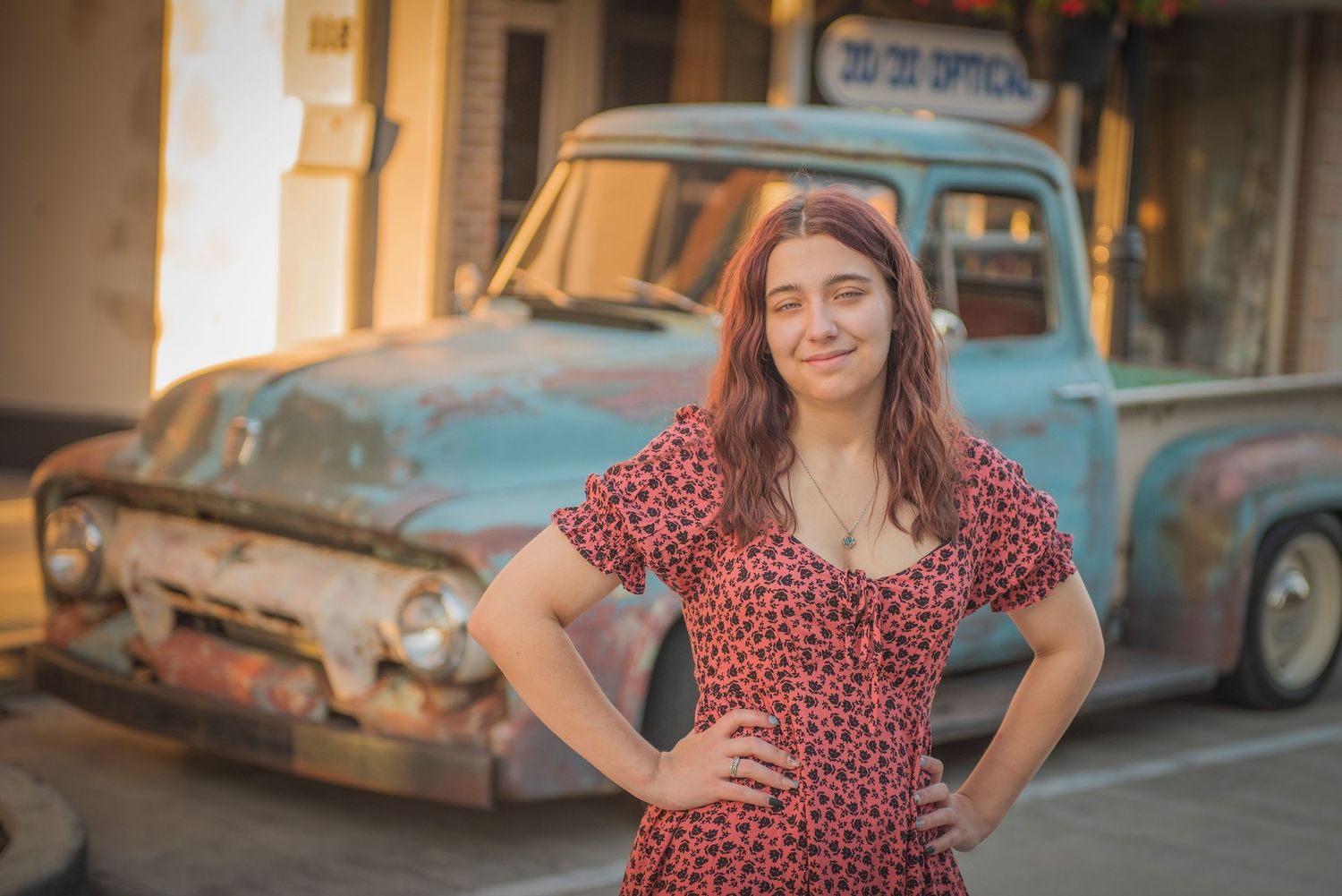 A person in a red polka dot dress poses in front of a vintage blue pickup truck during golden hour.