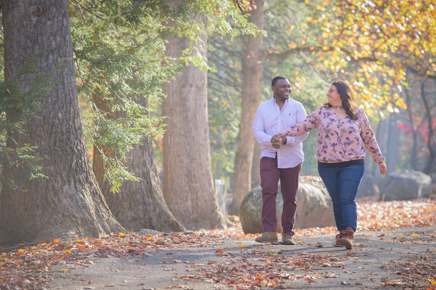 A couple walks hand in hand on a leaf-covered path through a park during autumn with golden foliage in the trees by Gatlinburg Photographers