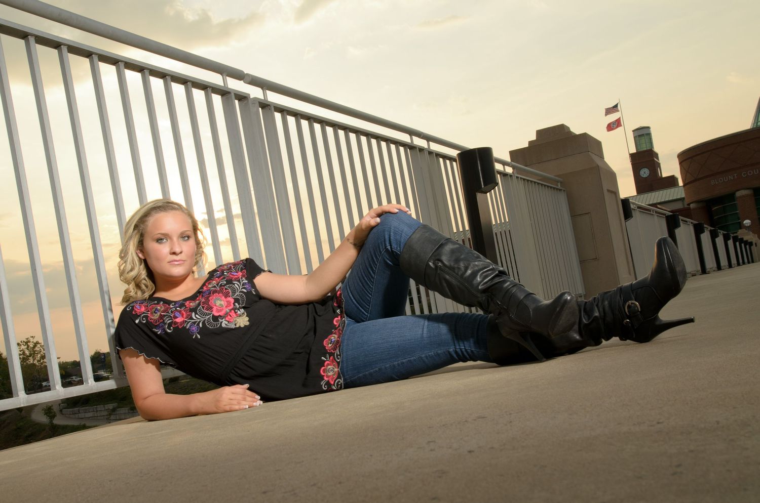 Someone in floral top and jeans poses casually on the ground against white railings during golden hour.