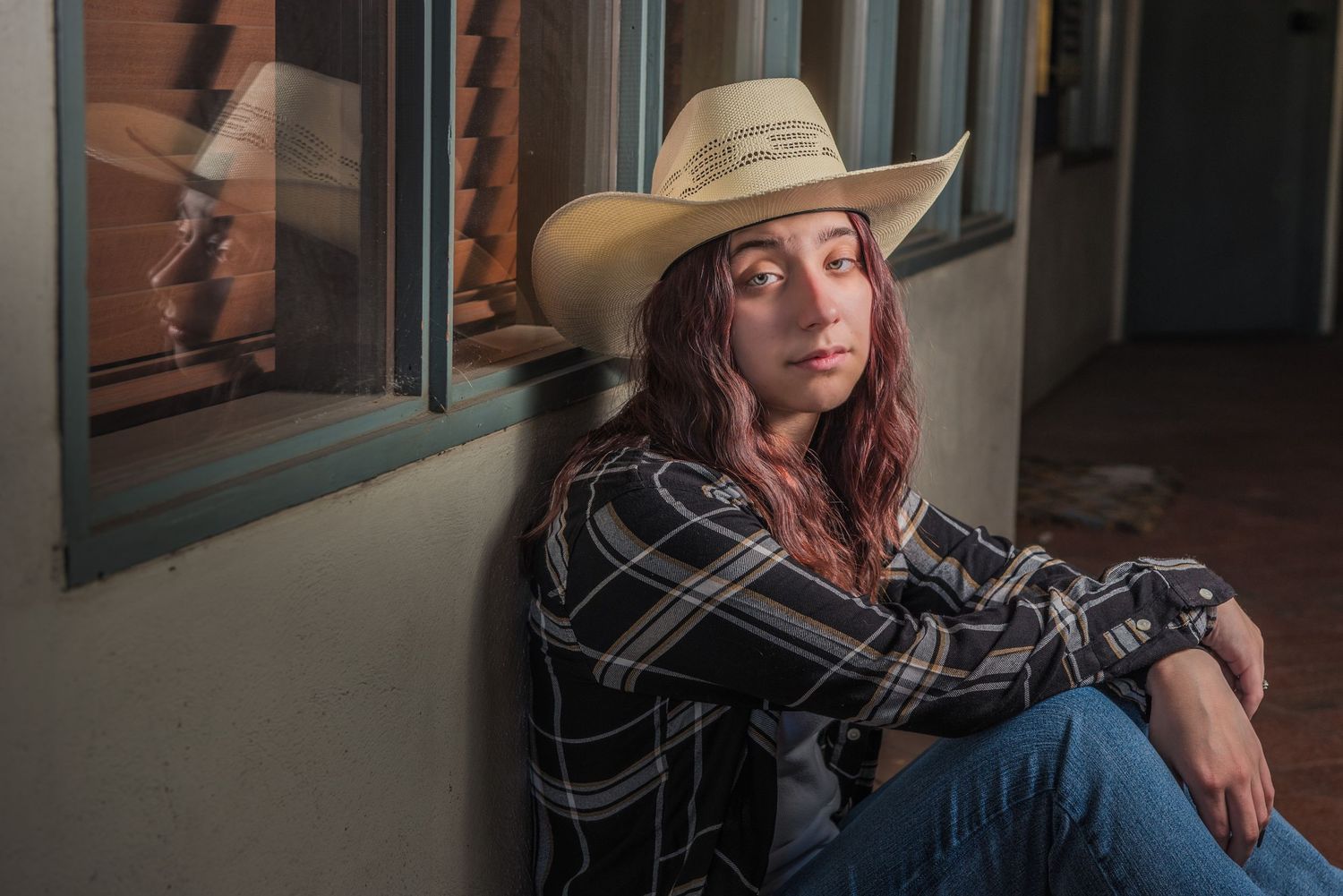 A person in a cowboy hat and plaid shirt sits casually against a wall in warm evening light.