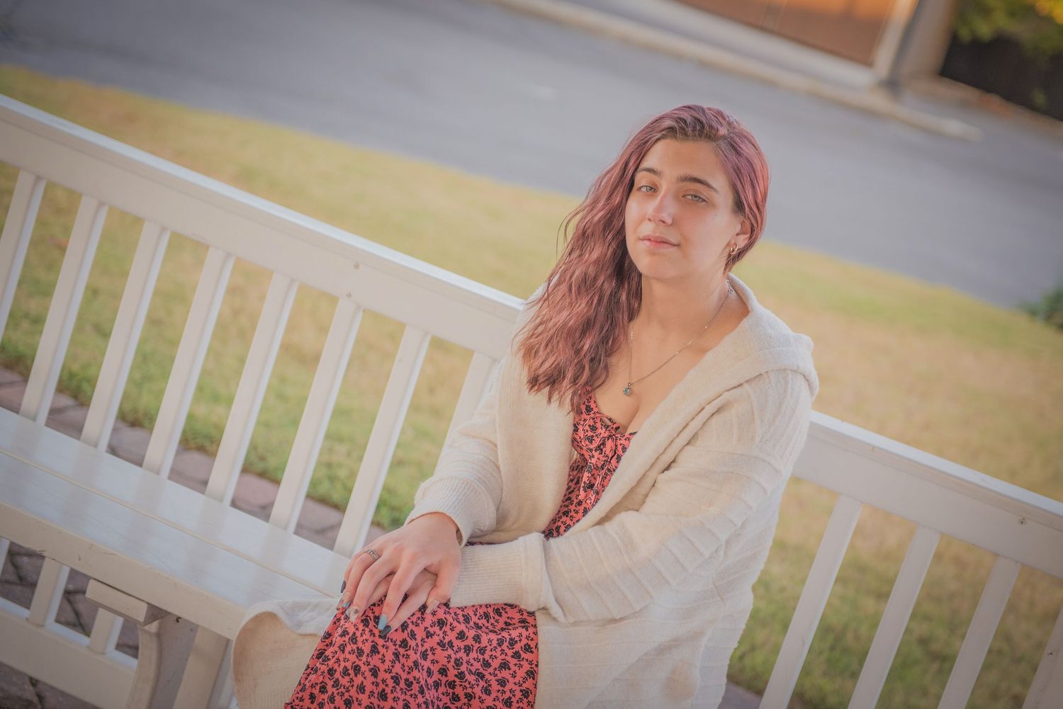 Someone wearing a cream cardigan and floral dress sits thoughtfully on a white wooden porch bench in warm sunlight.