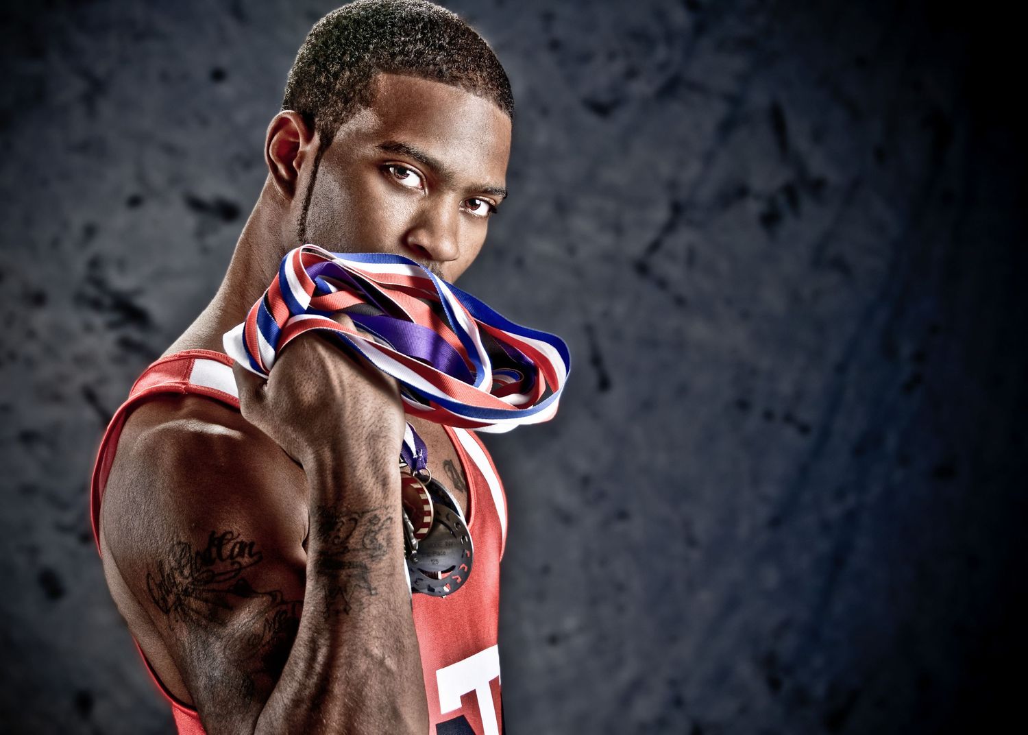 Athletic basketball player wearing a USA medal and red jersey poses against dark background showing bicep tattoo.