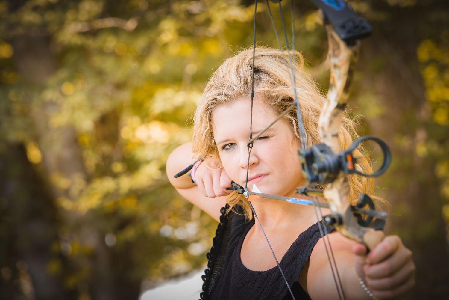 Archer in black tank top draws bow against autumn foliage background with golden bokeh lighting.
