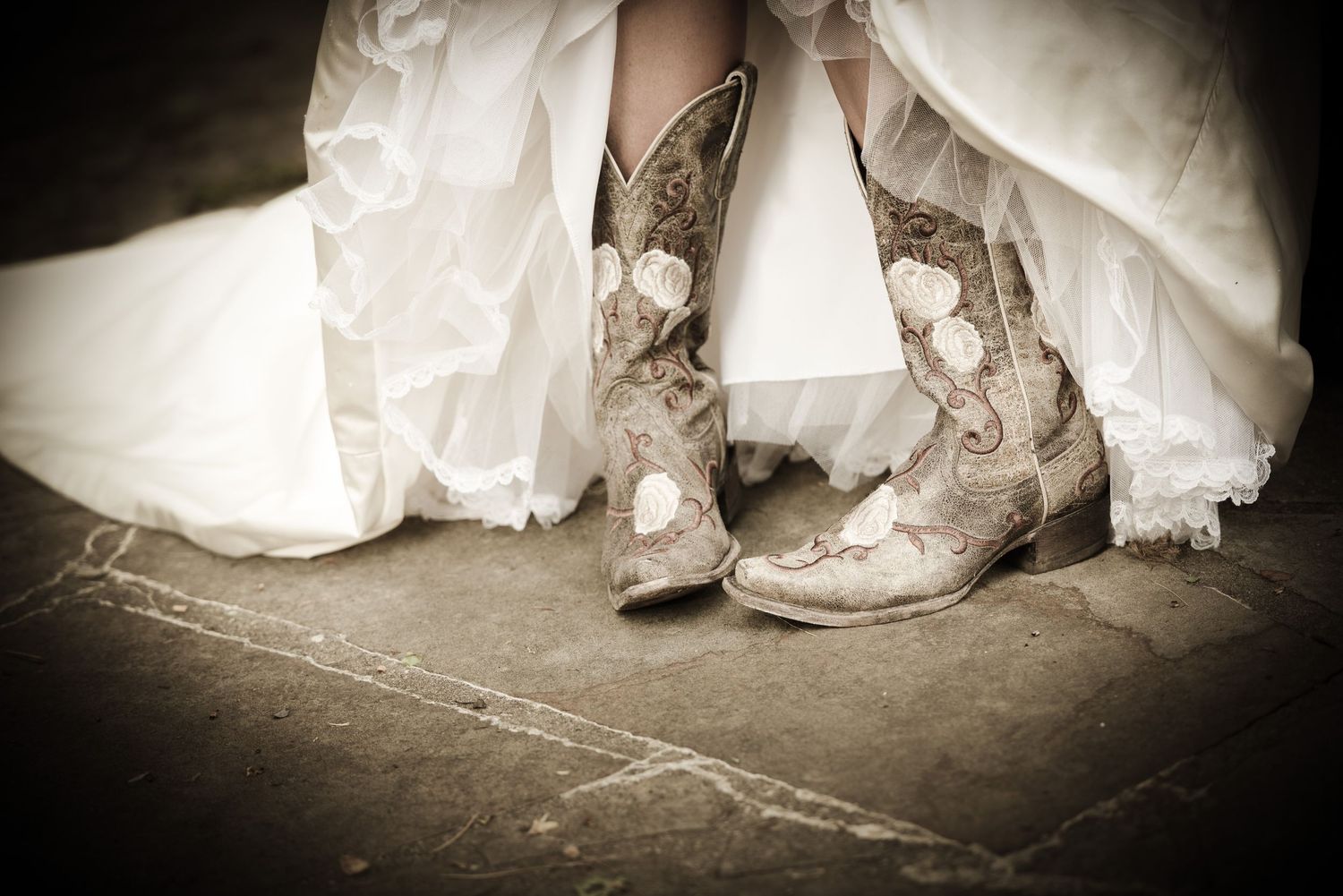 A pair of worn cowboy boots peek out from under a white wedding dress in a sepia-toned photograph by Gatlinburg Wedding Photographers.