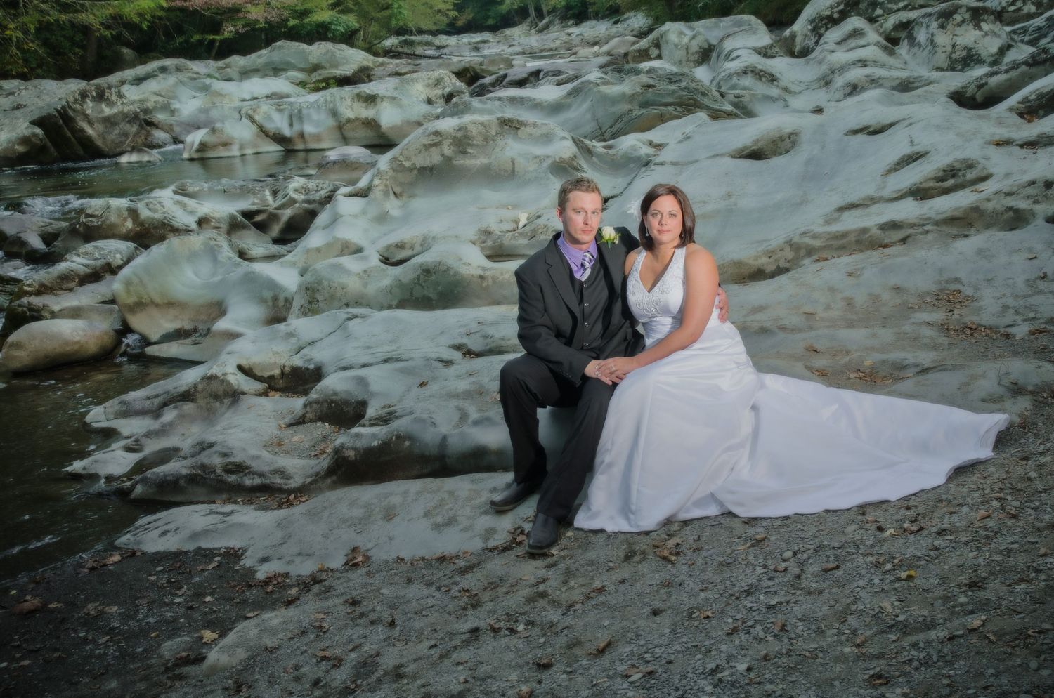 A couple in wedding attire sits together on a rocky beach during a coastal sunset photo session in Townsend TN.