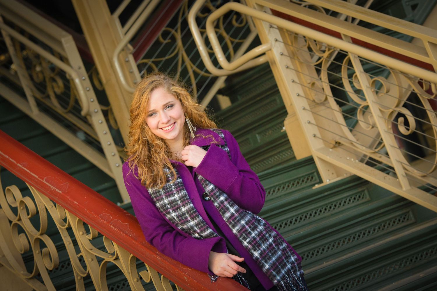 A person in a purple sweater and plaid dress sits on ornate red and gold vintage staircase railing.