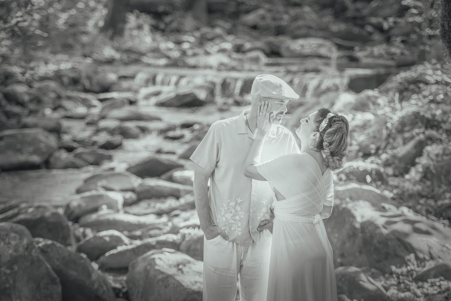 An anniversary couple stand together on rocky riverbank in romantic black and white photograph by Sevierville TN Photographers.