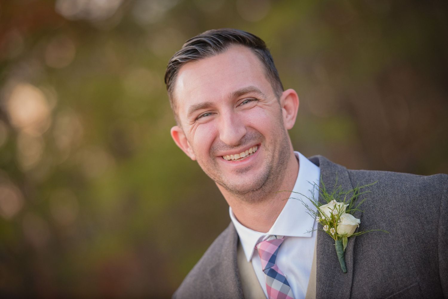 A smiling person in a grey suit with a boutonniere poses outdoors by Gatlinburg TN Wedding Photographers
