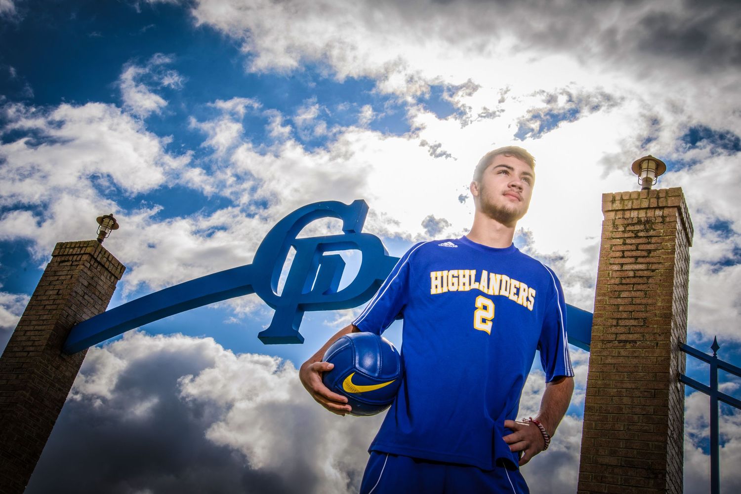 Athlete in blue jersey stands dramatically beneath cloudy sky with brick pillars and curved metal sports equipment.