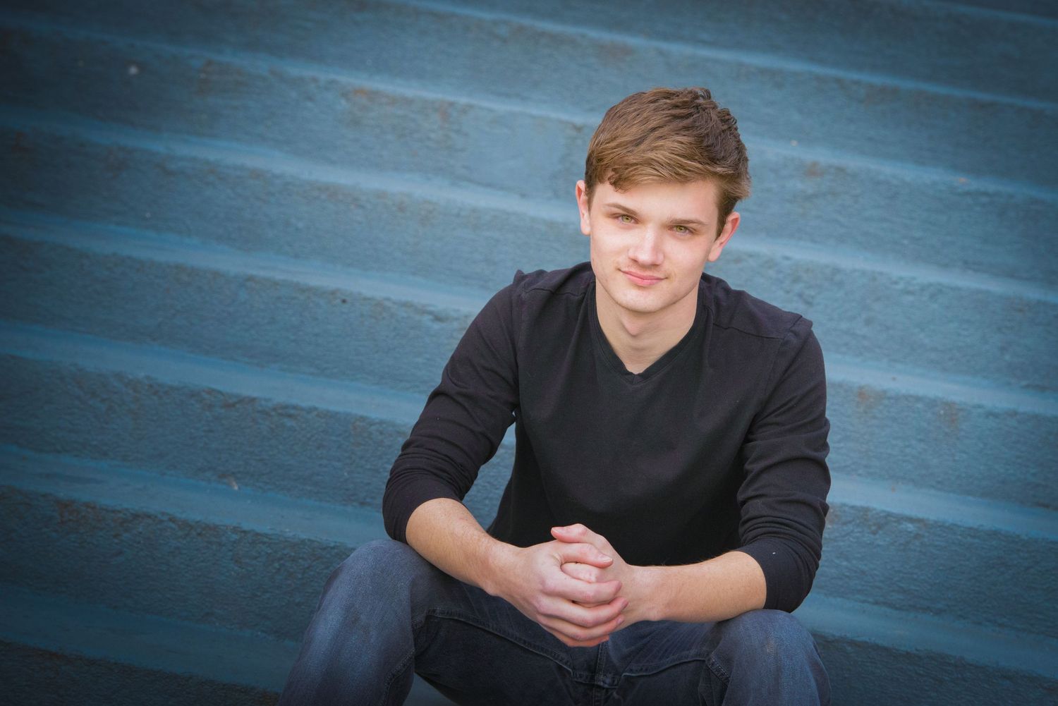 Subject in black v-neck sweater sits casually on blue steps with hands clasped during outdoor portrait session.