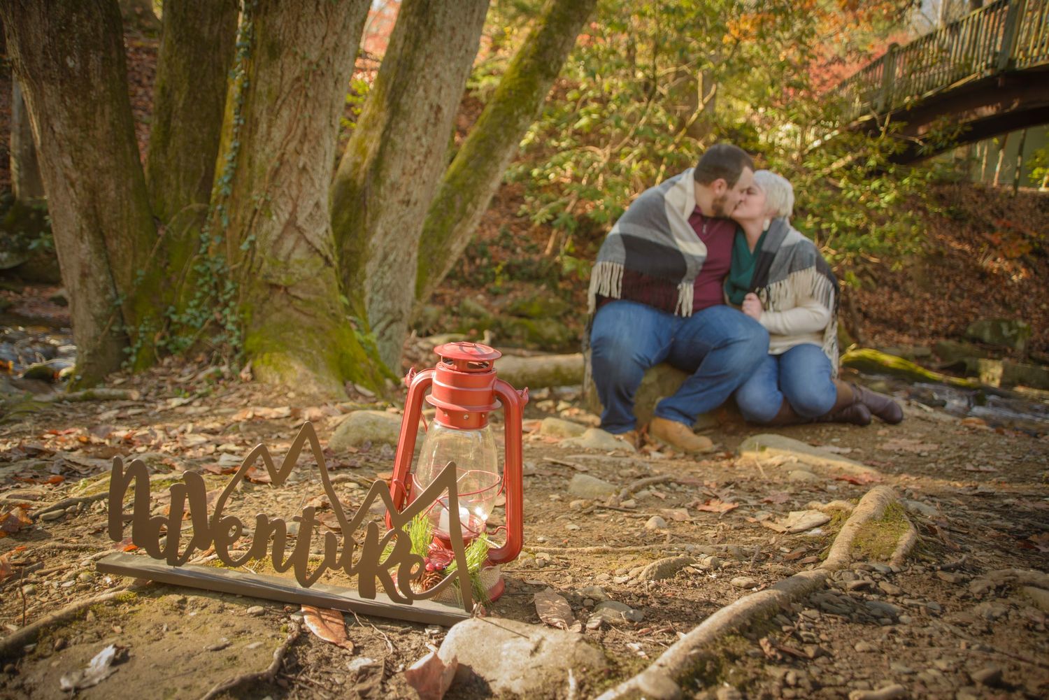 A couple sits together in autumn woods next to a rustic lantern and wooden Adventure sign in the fallen leaves by Sevierville Photographers.