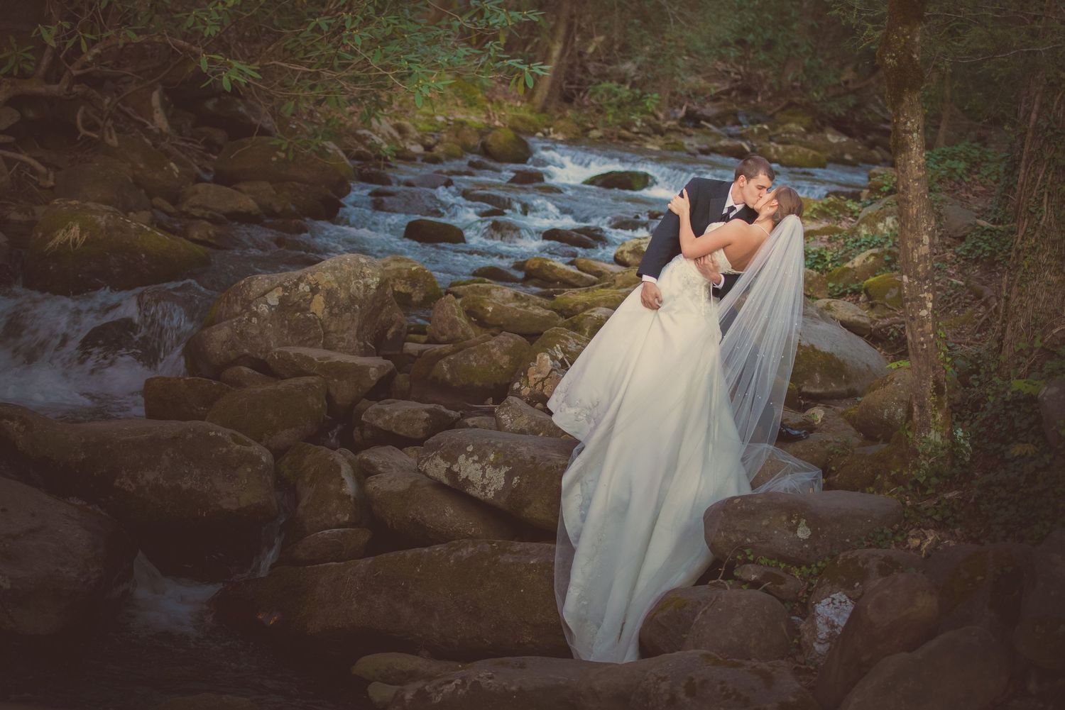 A flowing white wedding dress cascades over rocky terrain beside a rushing woodland stream in a romantic forest setting Mynatt Park.