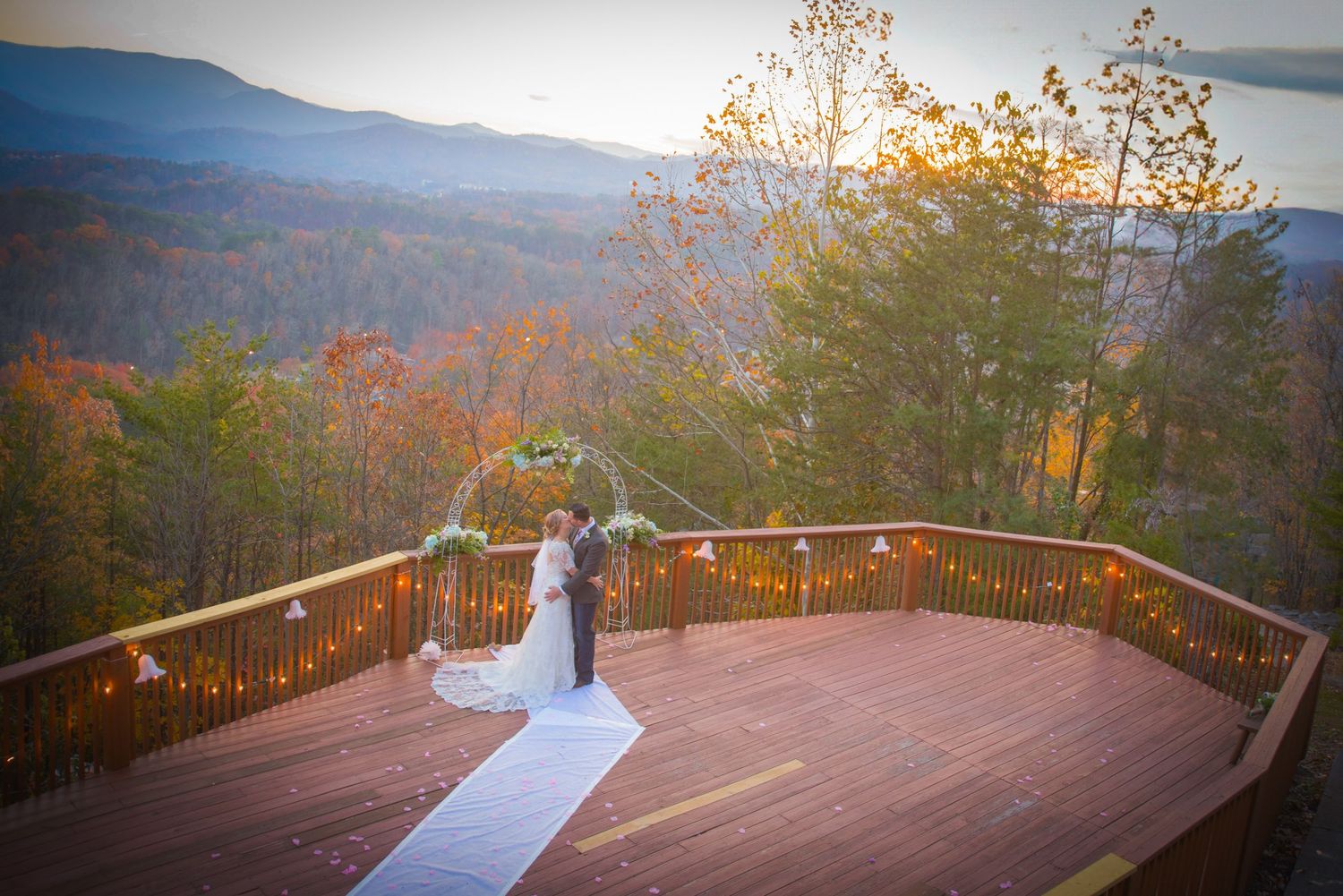 Two figures share a romantic dance on a wooden deck overlooking a mountain landscape at sunset during autumn.