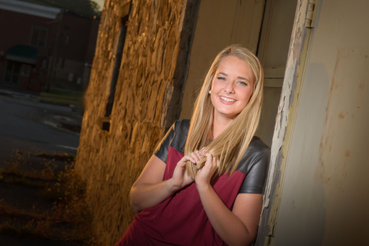 A person with long blonde hair wearing a burgundy shirt leans against a wooden post while posing indoors.