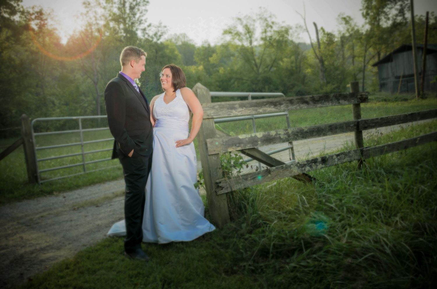 A couple poses on a country road at sunset with wooden fence and rustic barn in the background by Pigeon Forge Wedding Photographers.