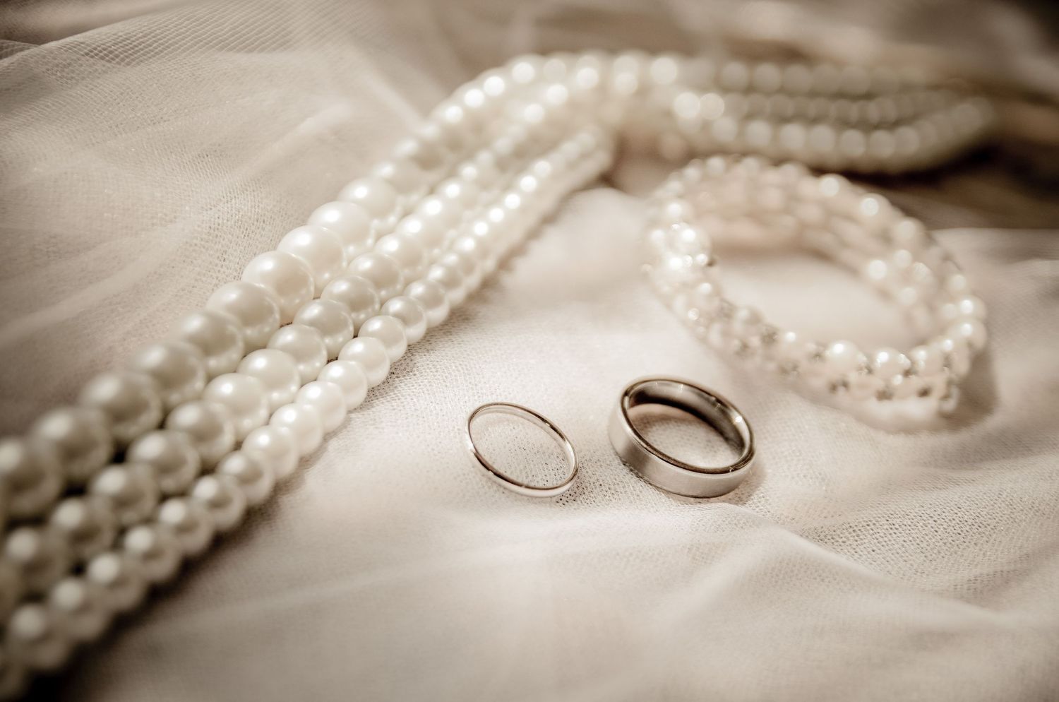 Two silver wedding rings rest beside strands of delicate white pearls on soft fabric in a vintage style composition Sevierville TN.