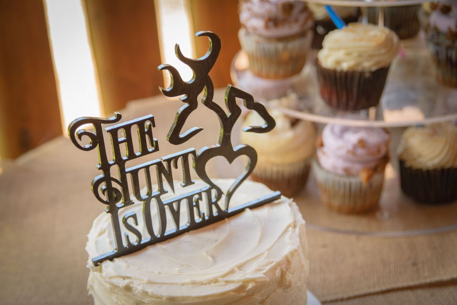 A rustic wedding cake topper reading 'The Hunt Is Over' with an antler design, displayed in front of out-of-focus cupcakes in Pigeon Forge.