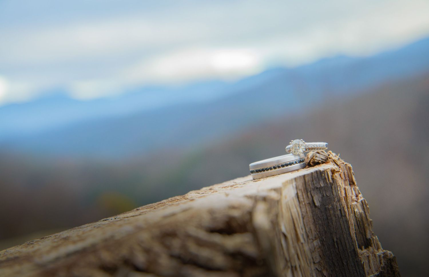 Two wedding rings sit on a weathered wooden surface with blurred mountaintops in the background by Gatlinburg Wedding Photographers.