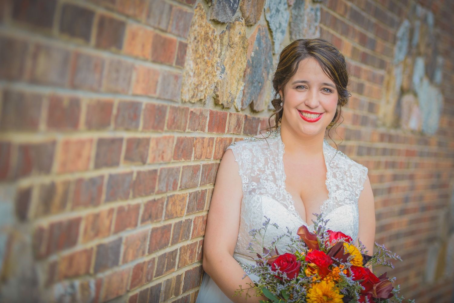 A bride poses with a colorful autumn bouquet of red and orange flowers against a rustic brick wall by Townsend Wedding Photographers.