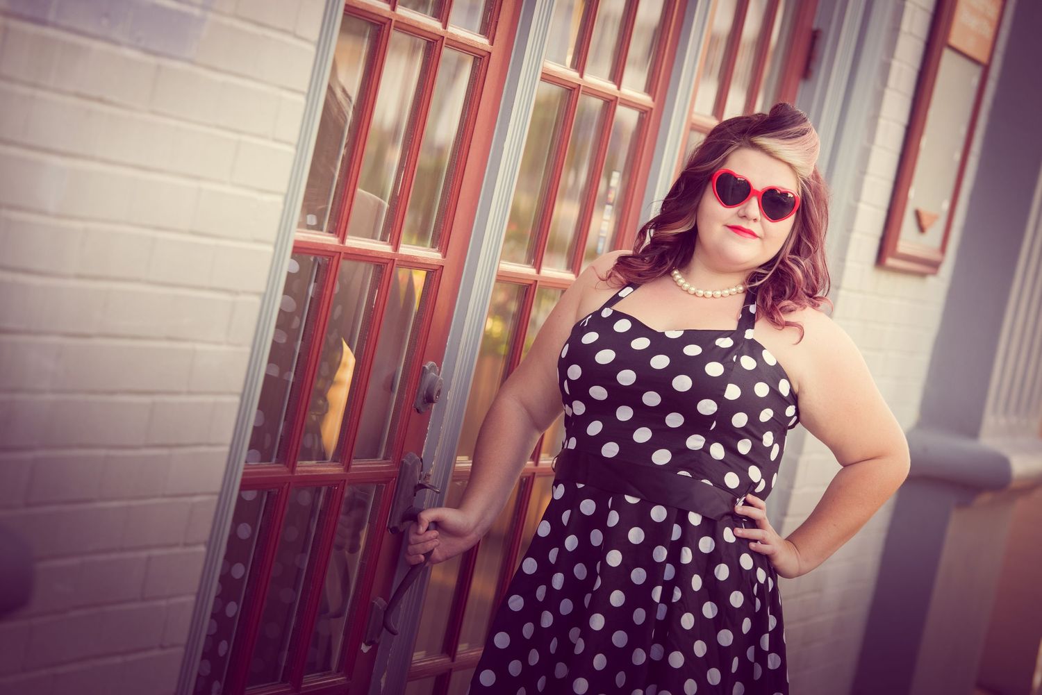A retro-styled photo of someone in a polka dot dress and sunglasses posing outdoors against a building wall.