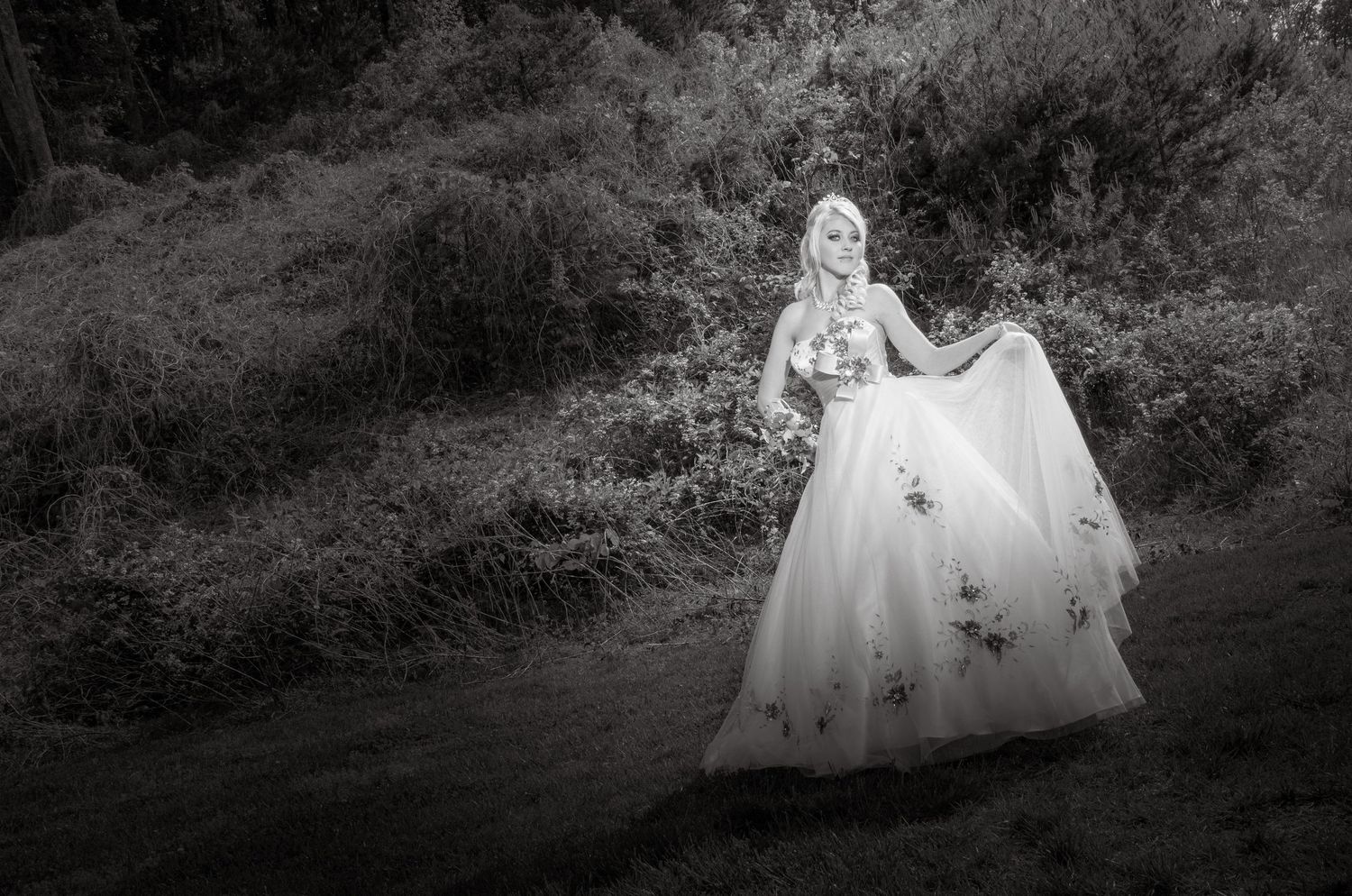 A black and white artistic portrait of a person in an elegant floral ballgown standing in a dreamy woodland setting.