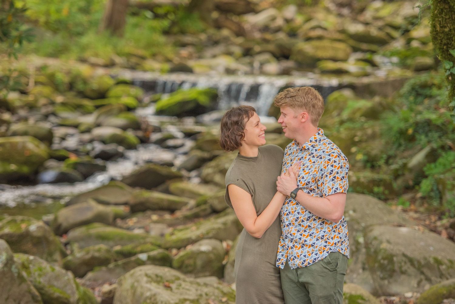 An anniversary couple embraces near a scenic waterfall with moss-covered rocks in a peaceful forest setting by Gatlinburg TN Photographer.