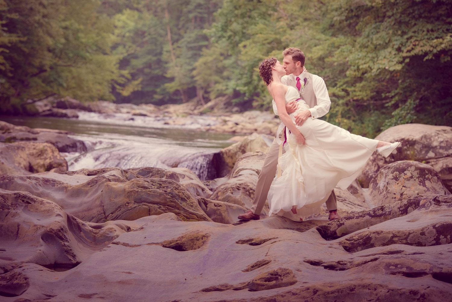 A romantic couple in formal attire share a tender moment on rocky riverbank with flowing water and forest backdrop by Gatlinburg Weddings.