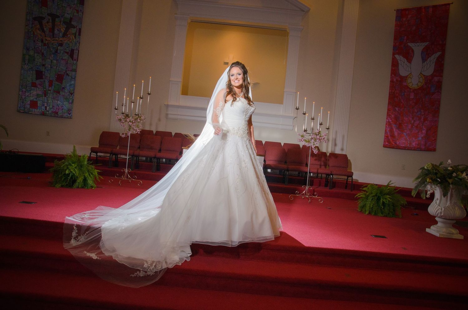 A bride in a flowing white wedding gown and veil poses in a church sanctuary with candleabras and red carpet in Sevierville TN.