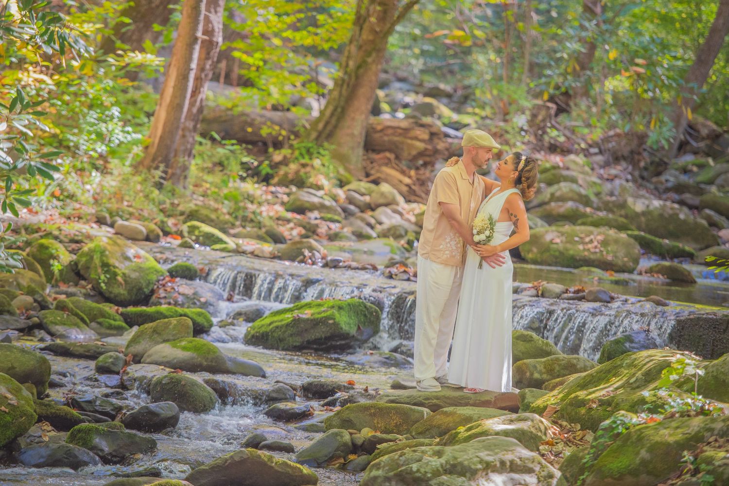 Two people embrace beside a tranquil forest stream with mossy rocks and cascading water on a sunlit summer day by Gatlinburg Photographers.