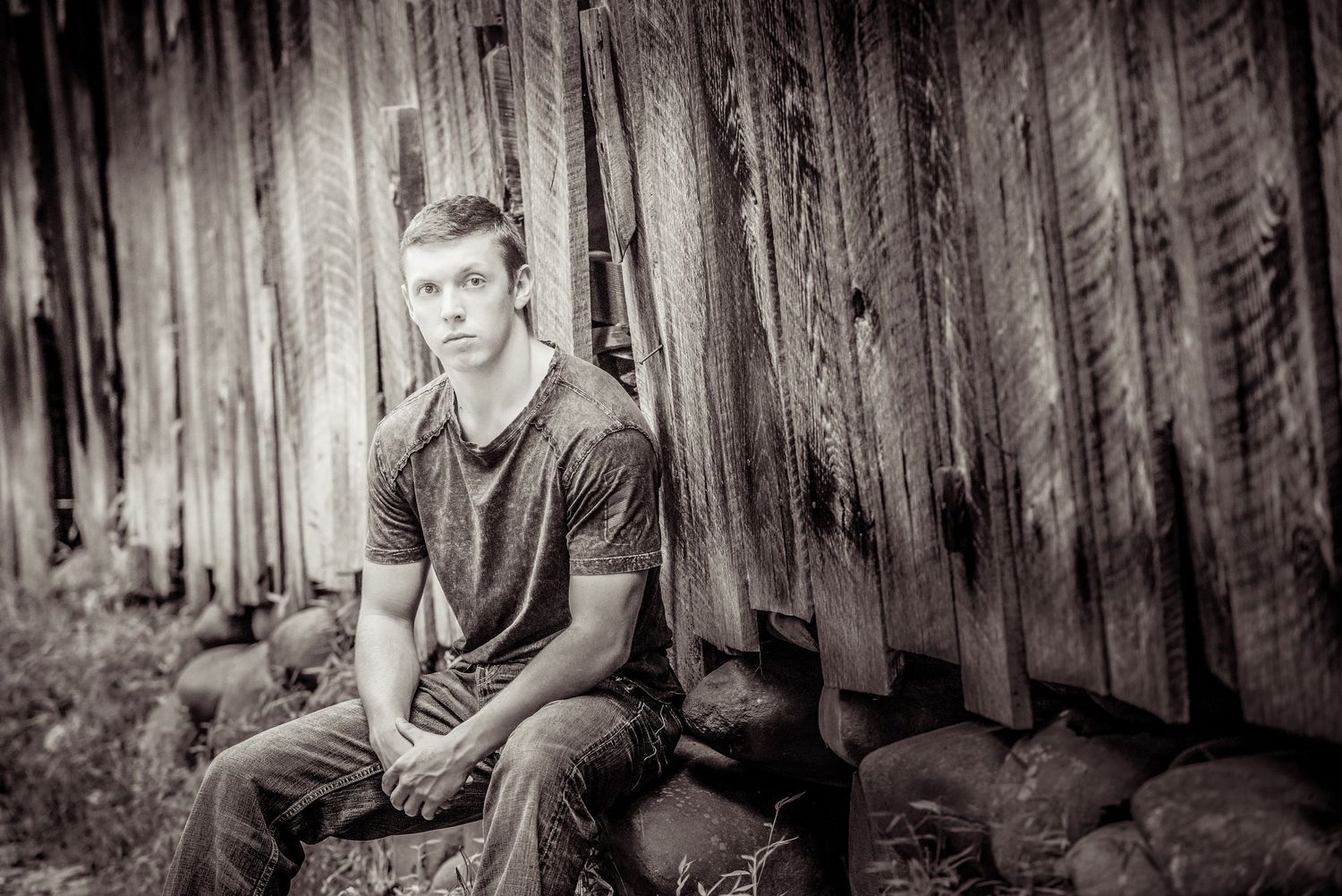 Black and white portrait of a young person seated on rocks against a rustic wooden wall backdrop.