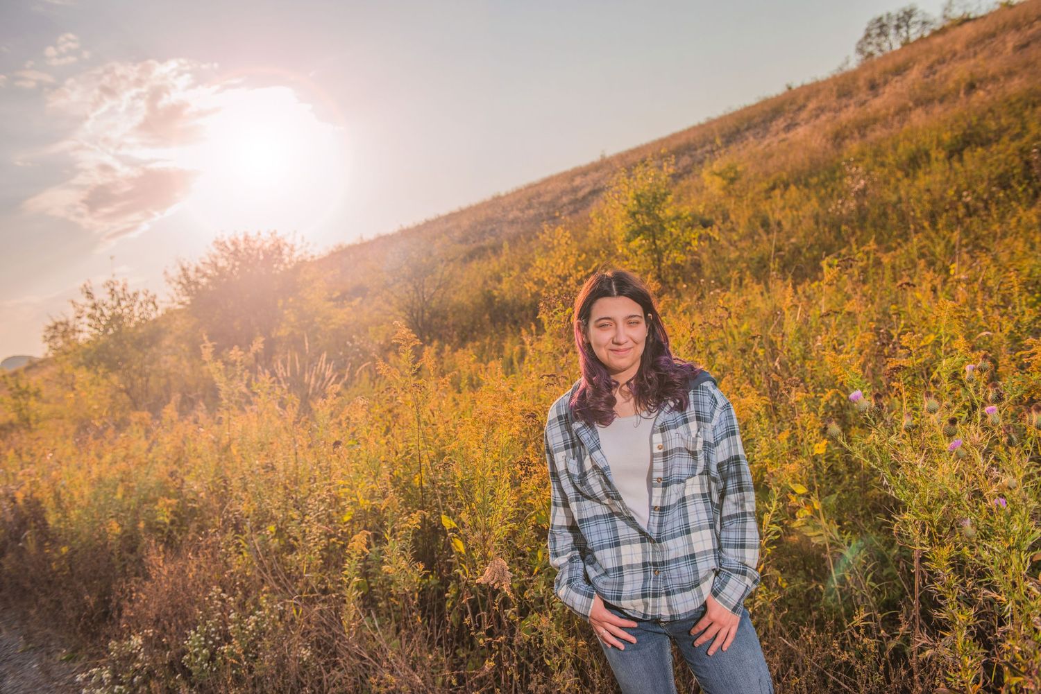 Someone stands on a golden hillside wearing a plaid shirt at sunset as warm light filters through autumn foliage.