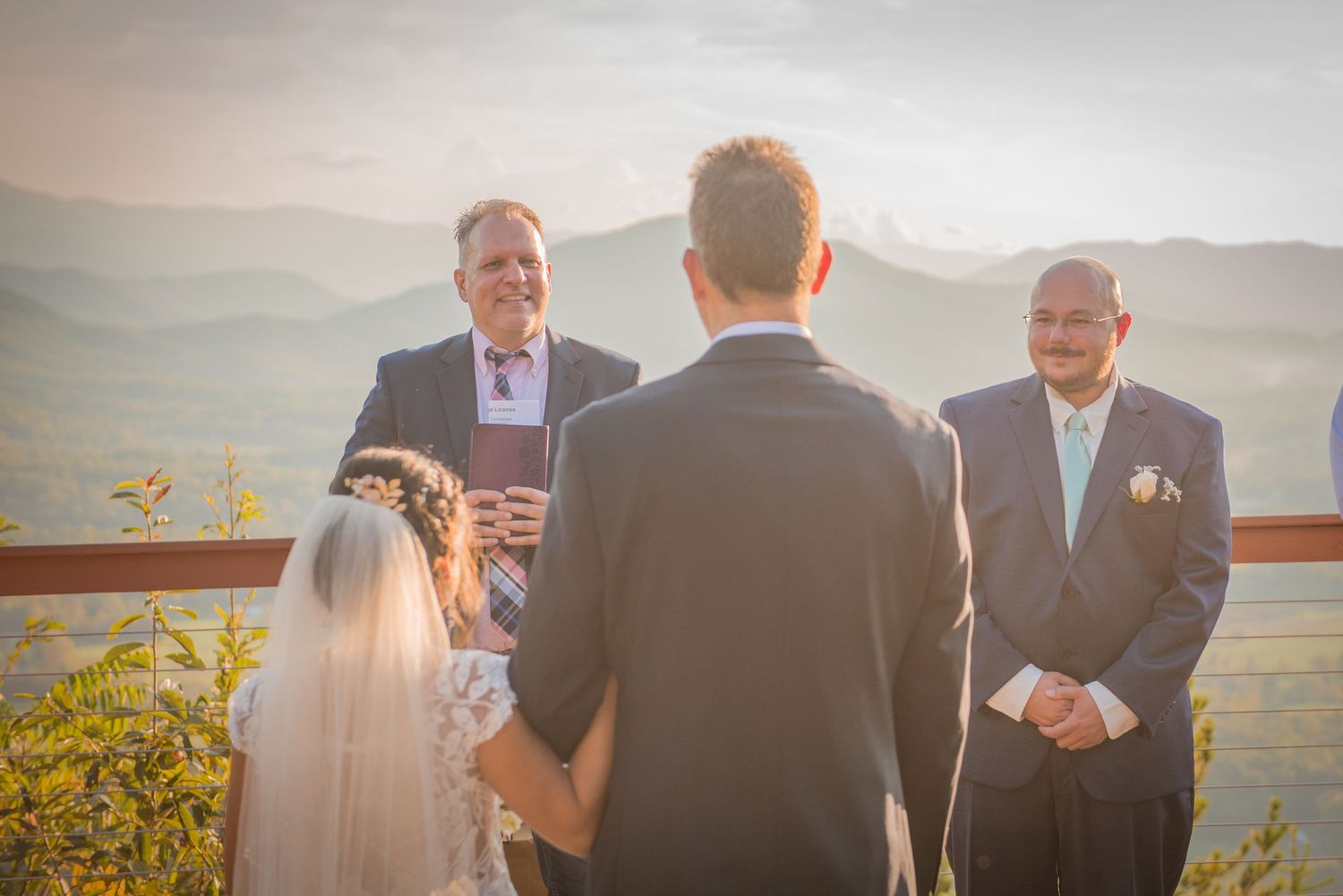 An outdoor wedding ceremony takes place on a mountaintop terrace with scenic views of rolling hills at sunset.