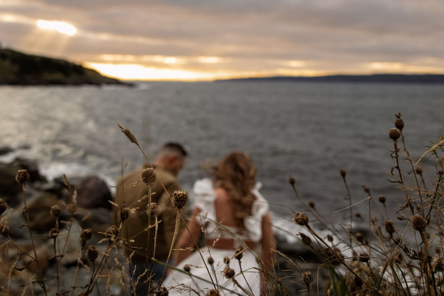 A couple walks on rocky coastline at sunset as waves crash against the shore and dried plants sway in foreground.