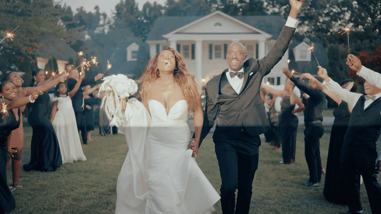 A joyful newlywed couple exits their wedding celebration while guests wave and cheer on the lawn of a white columned venue.
